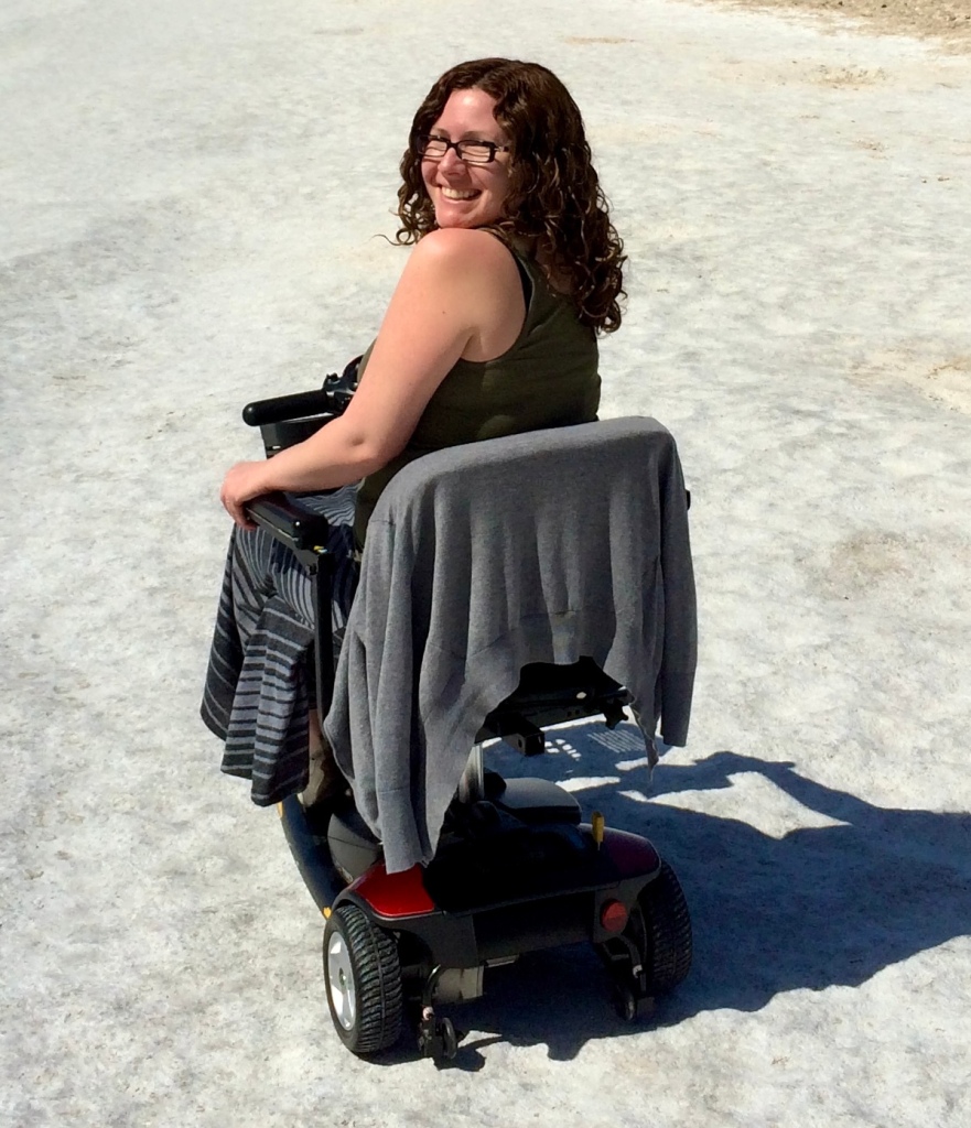 Suzanne is sitting in her mobility scooter looking back over her shoulder. She is wearing a tank top and a long skirt. She has long curly hair and is wearing glasses. ￼